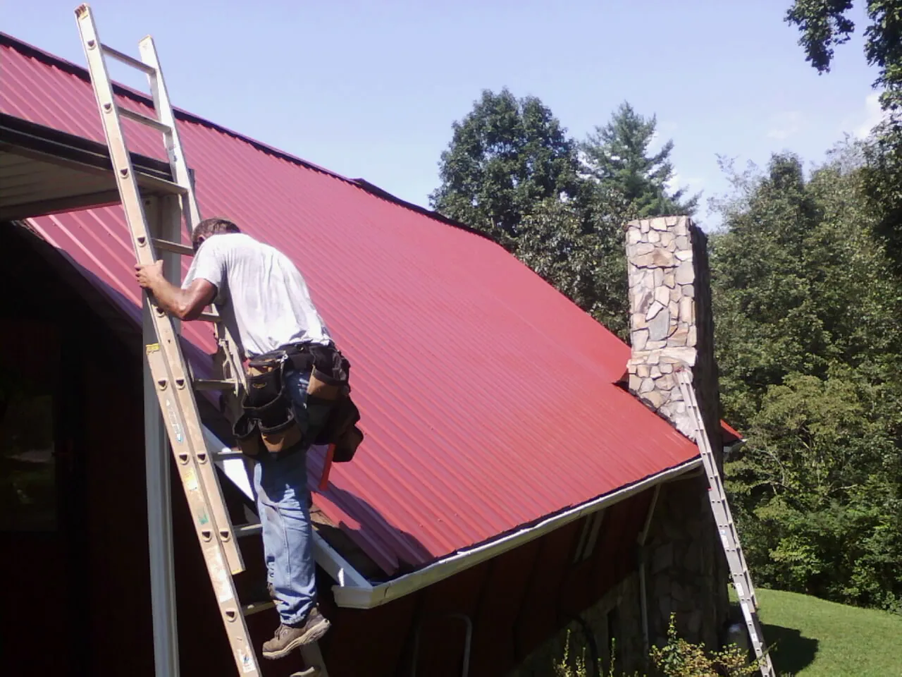 Roofer on a ladder beside a red metal roof and stone chimney — Soffit & Fascia Repair by Roofing Brothers in Omaha